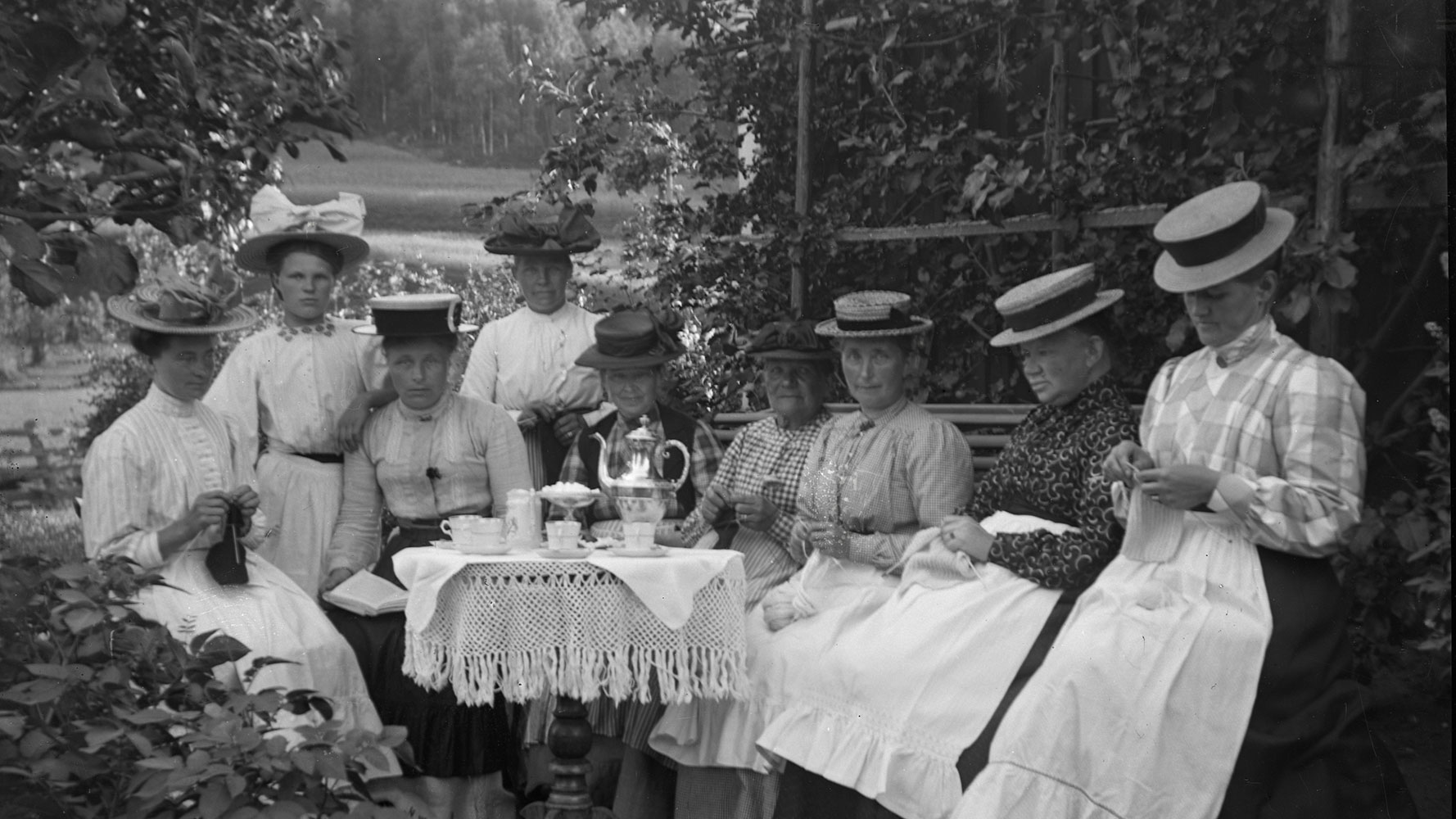 Svartvitt gammalt fotografi. En samling kvinnor i gammaldags klänningar och hattar poserar för porträtt ute i trädgården. De är samlade runt bord med vita spetsdukar och har handarbeten i sina händer.