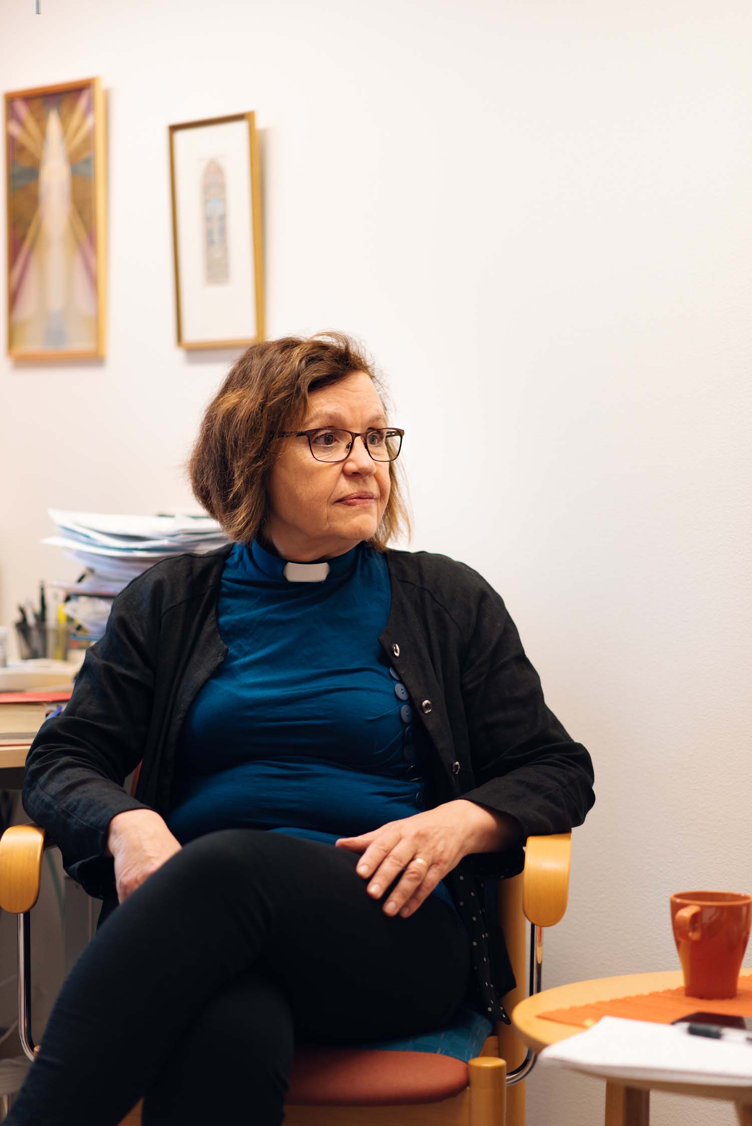 En kvinna med prästkrage sitter i en fåtölj och tittar åt höger ut bild. Hon har en blå tröja och halvkort, brunt hår.