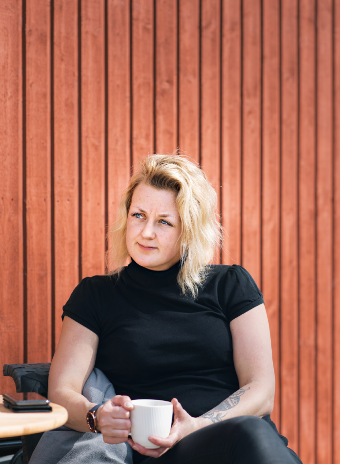 En kvinna med blont hår sitter framför en faluröd trävägg. Hon bär en svart vt-shirt och håller i en vit kaffekopp.,