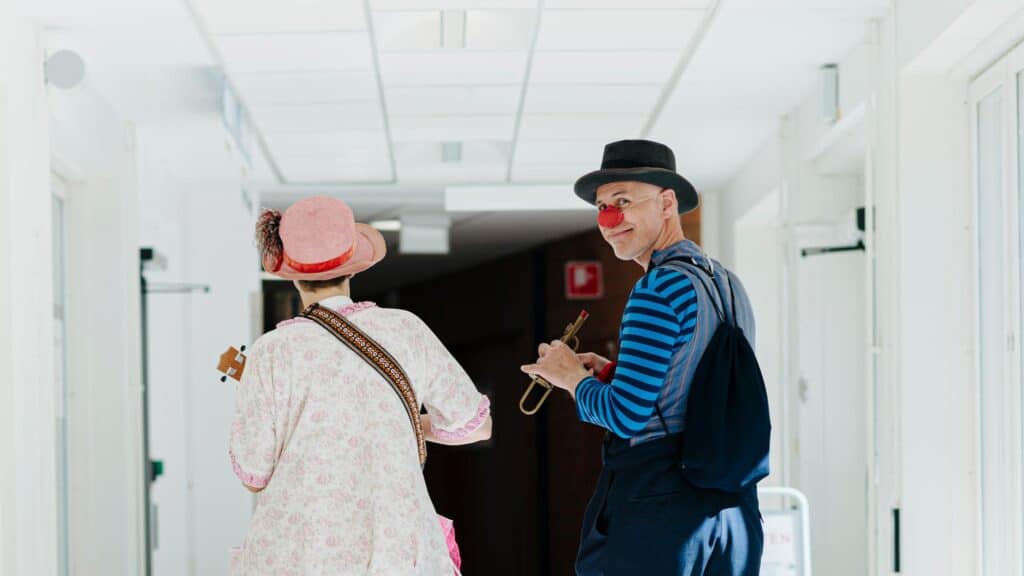 Två sjukhusclowner går i en sjukhuskorridor. Vi ser clownerna bakifrån. En av clownerna bär en trumpet och tittar bakåt mot kameran.